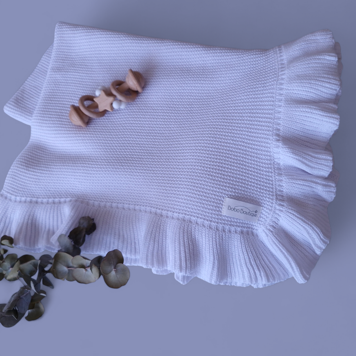 A neatly folded Ruffled Blanket from BOBO BOUTIQUE, crafted in soft white knit with elegant ruffled edges, features a customizable label on the bottom right. A wooden baby rattle is placed on top, while lush greenery enhances the cozy allure of these personalized baby blankets.