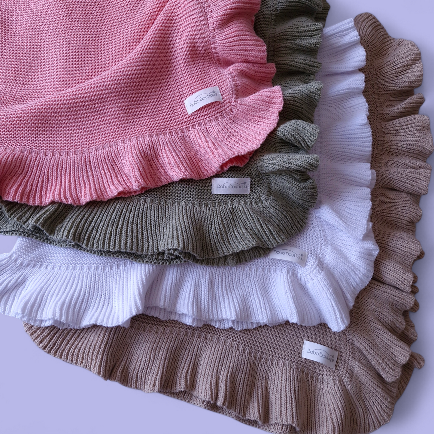 Four Ruffled Blankets by BOBO BOUTIQUE are stacked on a flat surface, each featuring ruffled edges in pink, olive green, white, and beige. Small labels adorn the corners of these cozy creations. The light lavender background accentuates their charm.