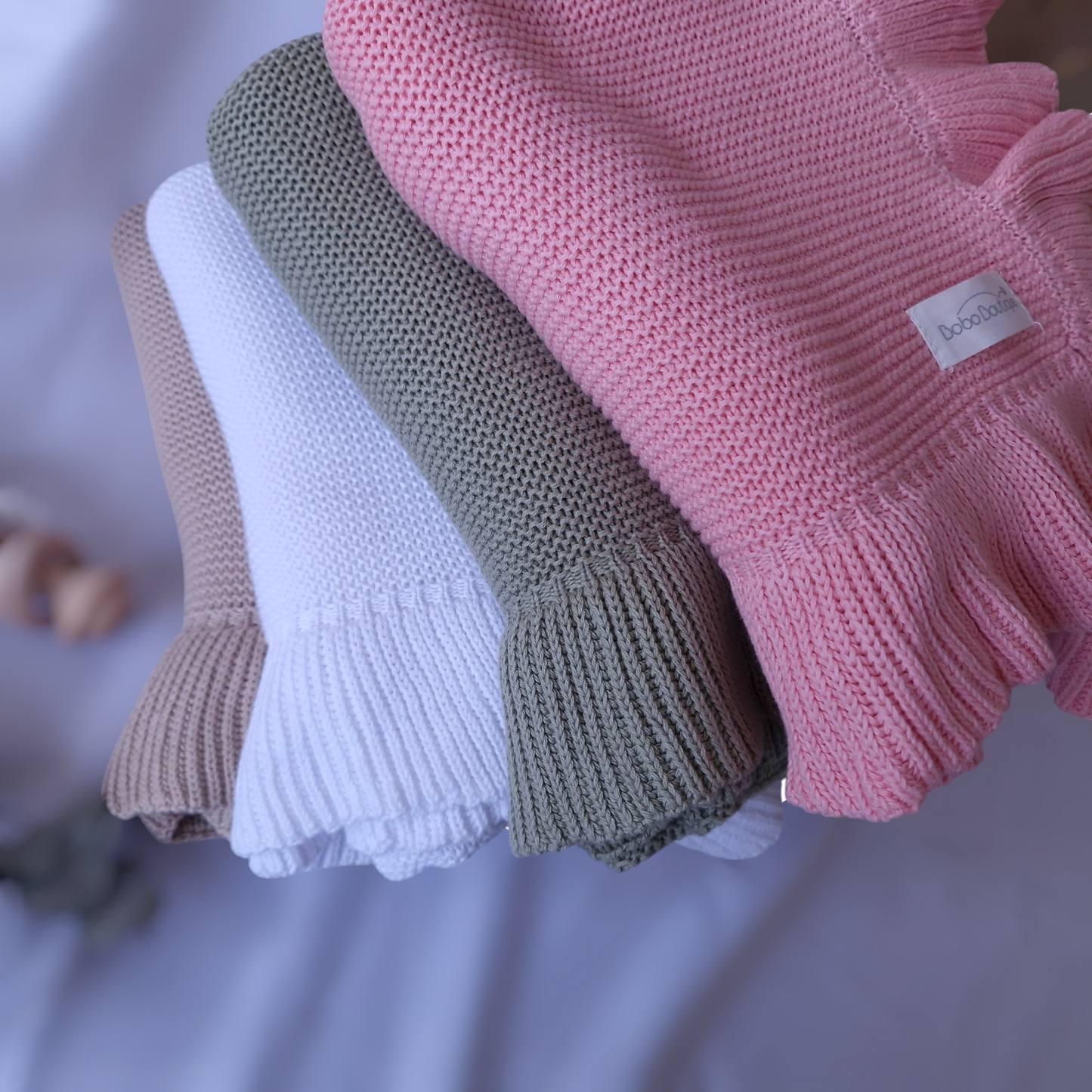 A collection of BOBO BOUTIQUE's Ruffled Blankets is neatly arranged in a spectrum of colors: pink, green, white, and beige. Each soft knitted piece, including the pink one with a visible label, is beautifully folded against a softly blurred background. These cozy throws offer the perfect opportunity to be customized as baby blankets with embroidery in your preferred color.